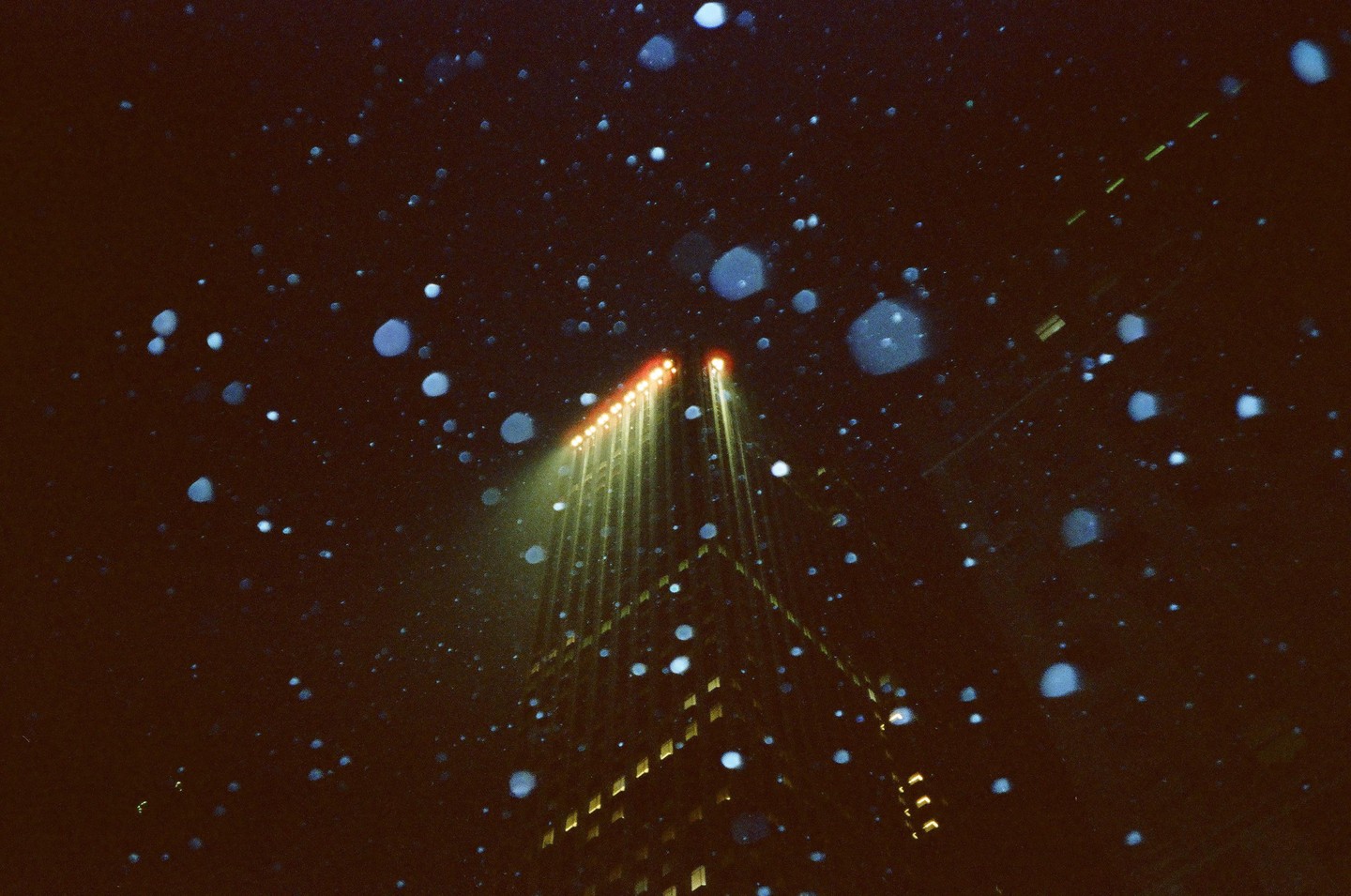 Snow falls around a skyscraper with two bright lights shining upwards into the night sky.