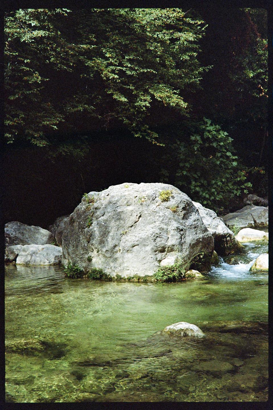 A large rock sits in a clear green river with trees in the background.