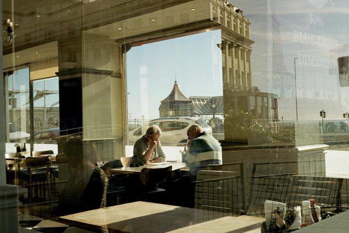 Two people sit at a table in a cafe with a view of a street and building through the window.