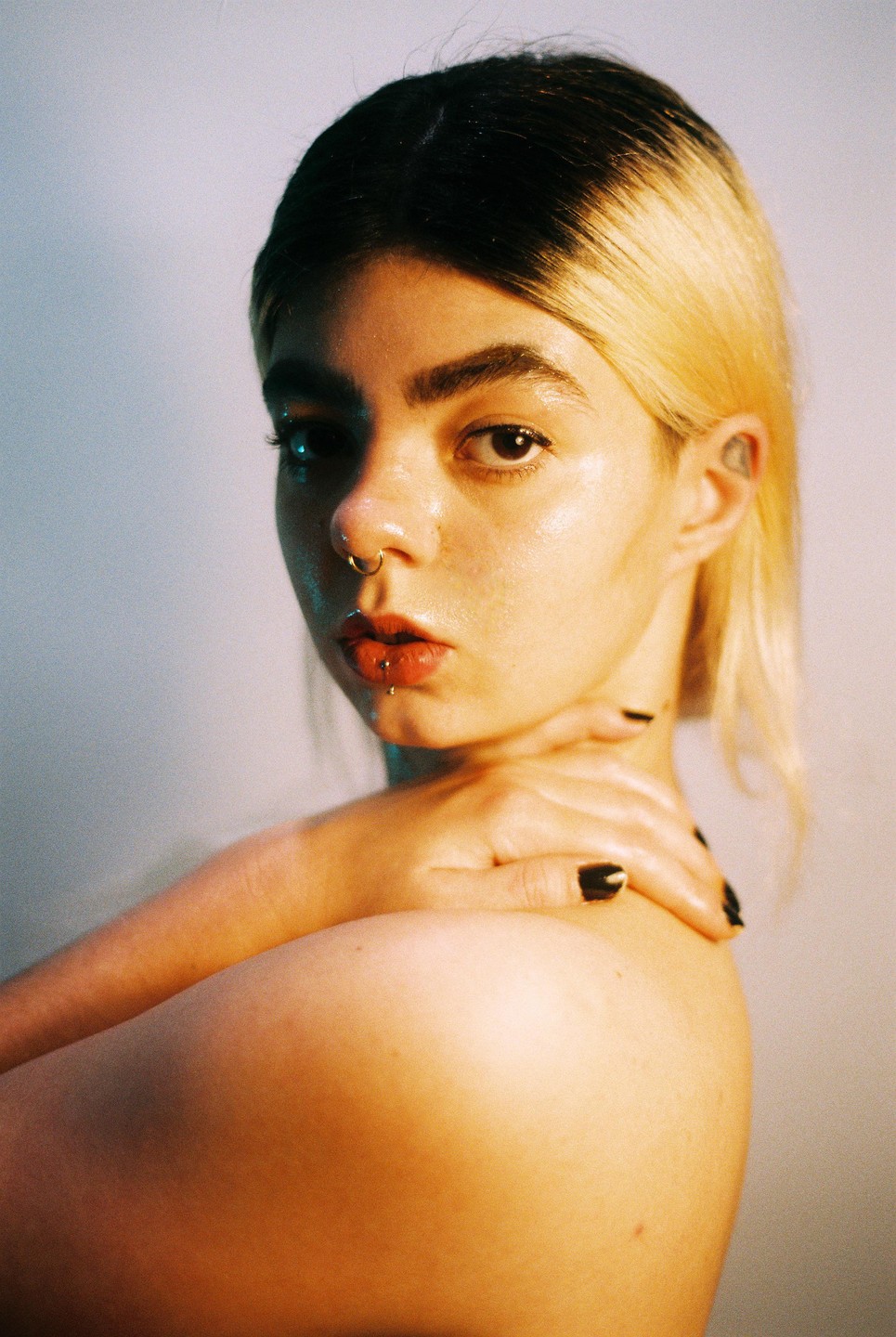 A woman with blonde hair and dark roots looks at the viewer, with her hands on her shoulder.