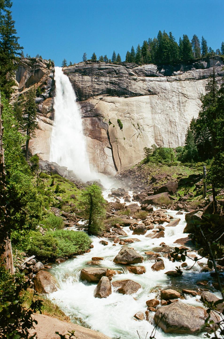 A large waterfall cascades down a rocky cliff into a river filled with boulders and surrounded by green trees.