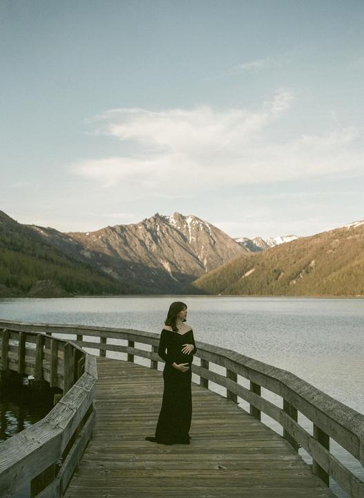 A pregnant woman stands on a wooden pier overlooking a lake surrounded by mountains.
