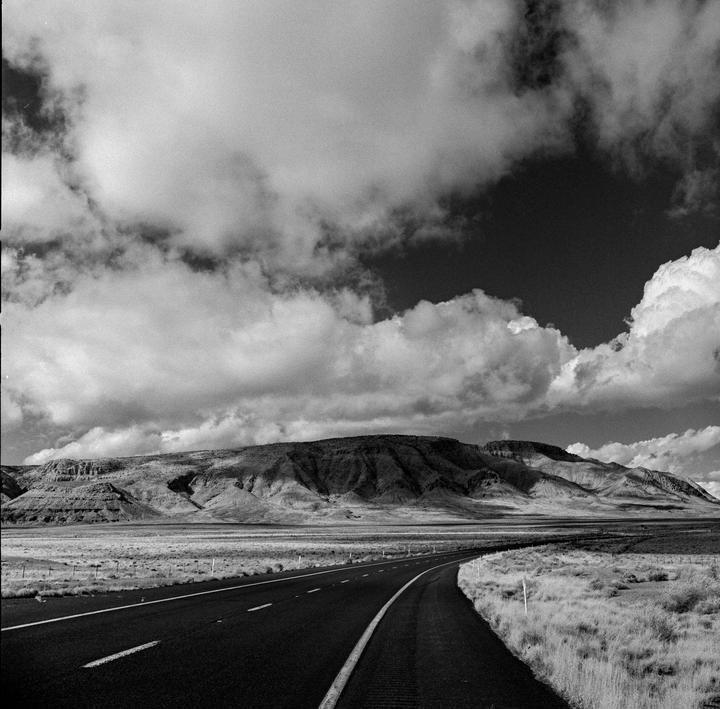 A long road curves through a vast, flat landscape towards a distant mountain range under a cloudy sky.