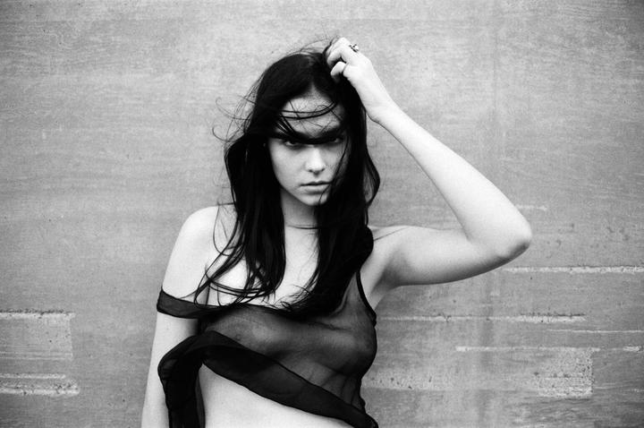 A woman with dark hair and a sheer top poses against a textured wall, with her hand in her hair.
