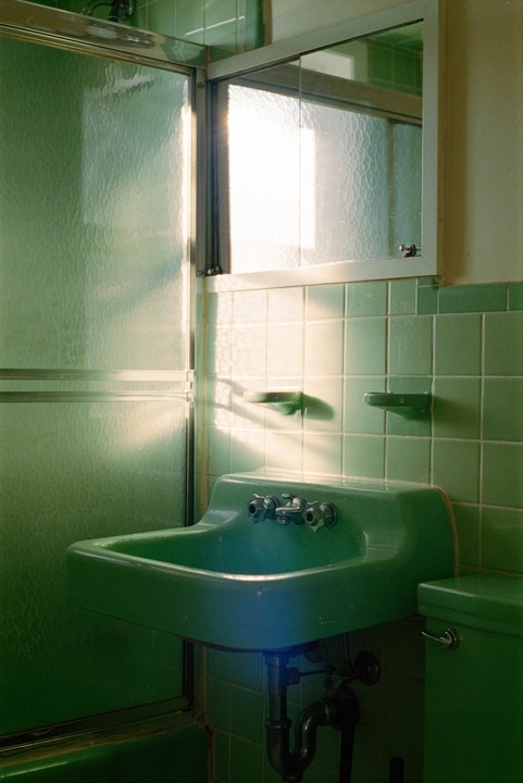 A green bathroom with a sink, shower, and mirror is illuminated by bright light from a window.