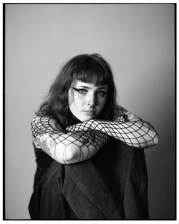 A woman with dark hair and bangs, wearing fishnet sleeves and jeans, looks directly at the viewer.