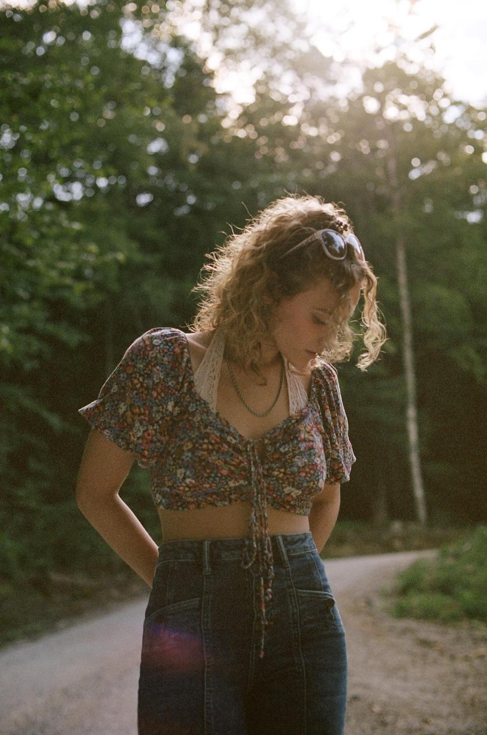 A woman with curly hair and sunglasses on her head stands on a dirt road, looking down.