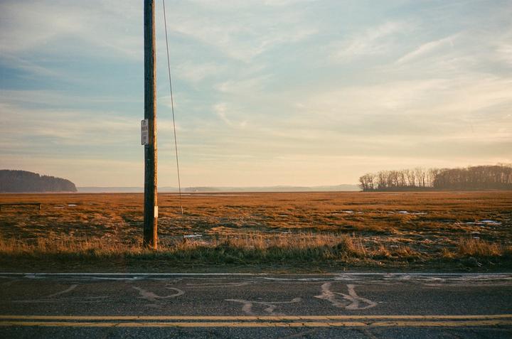 A utility pole stands beside a road with double yellow lines, overlooking a vast marshland under a partly cloudy sky.