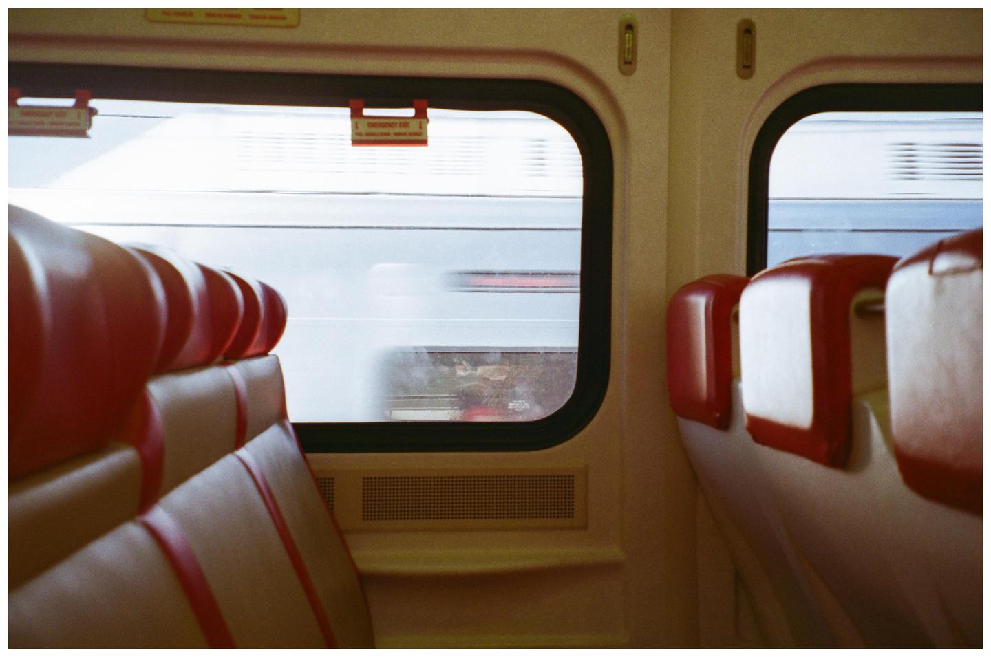 The interior of a train car with red and beige seats is visible, with a blurred view of another train passing by outside the window.