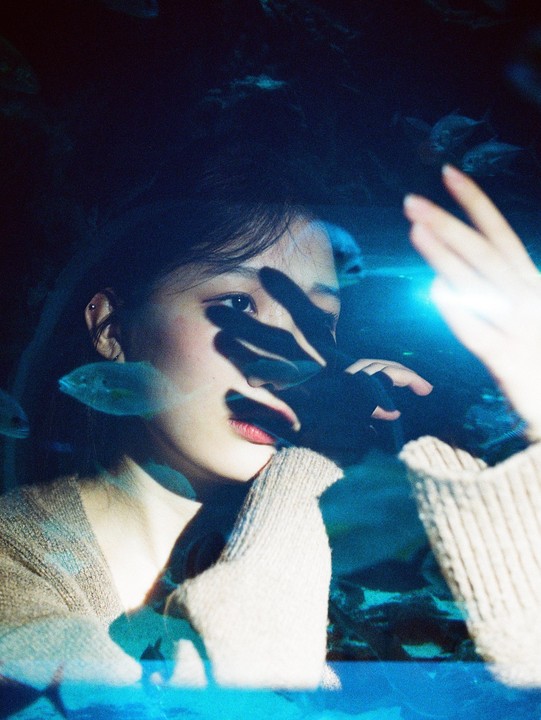 A woman with her hand in front of her face looks into an aquarium with fish swimming in blue water.