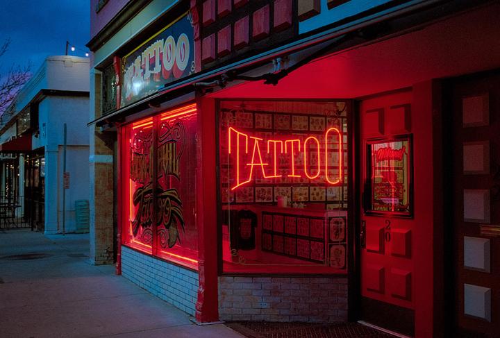 A tattoo shop with a red neon sign is visible from the sidewalk at dusk.