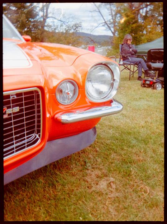 The front of an orange car with a chrome bumper is parked on grass, with a man sitting in a chair in the background.