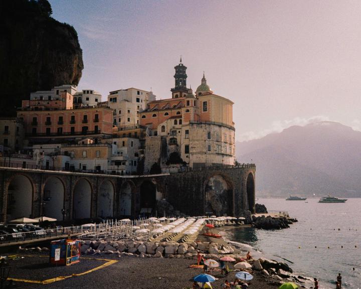 A coastal town with buildings built into a cliffside overlooks a beach with lounge chairs and umbrellas.