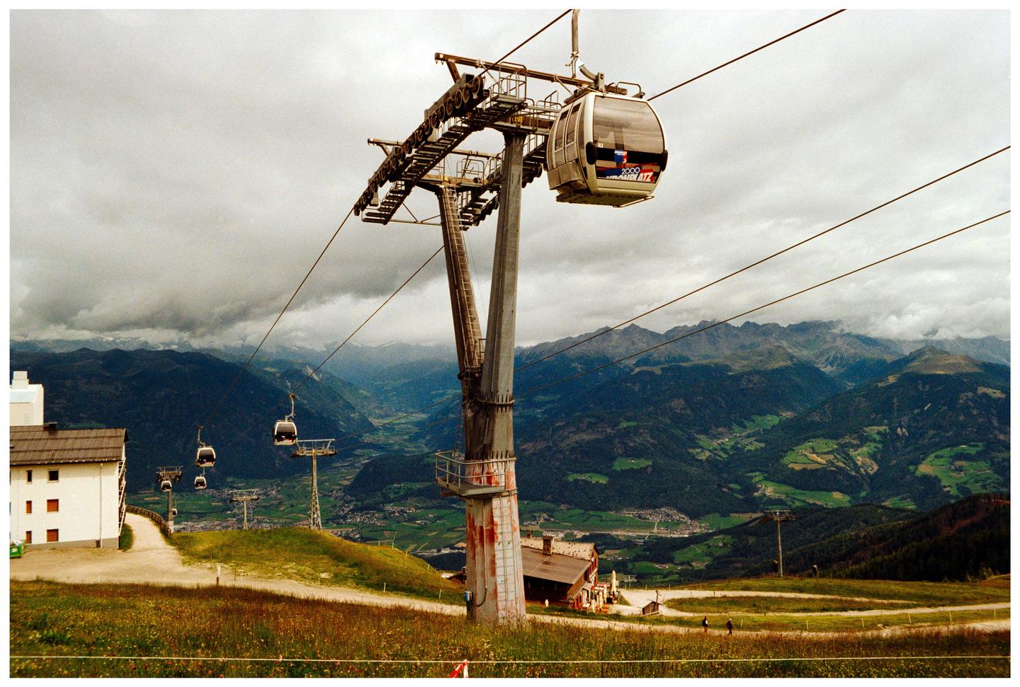 A gondola lift with several cabins is visible on a cloudy day, with mountains and a valley in the background.