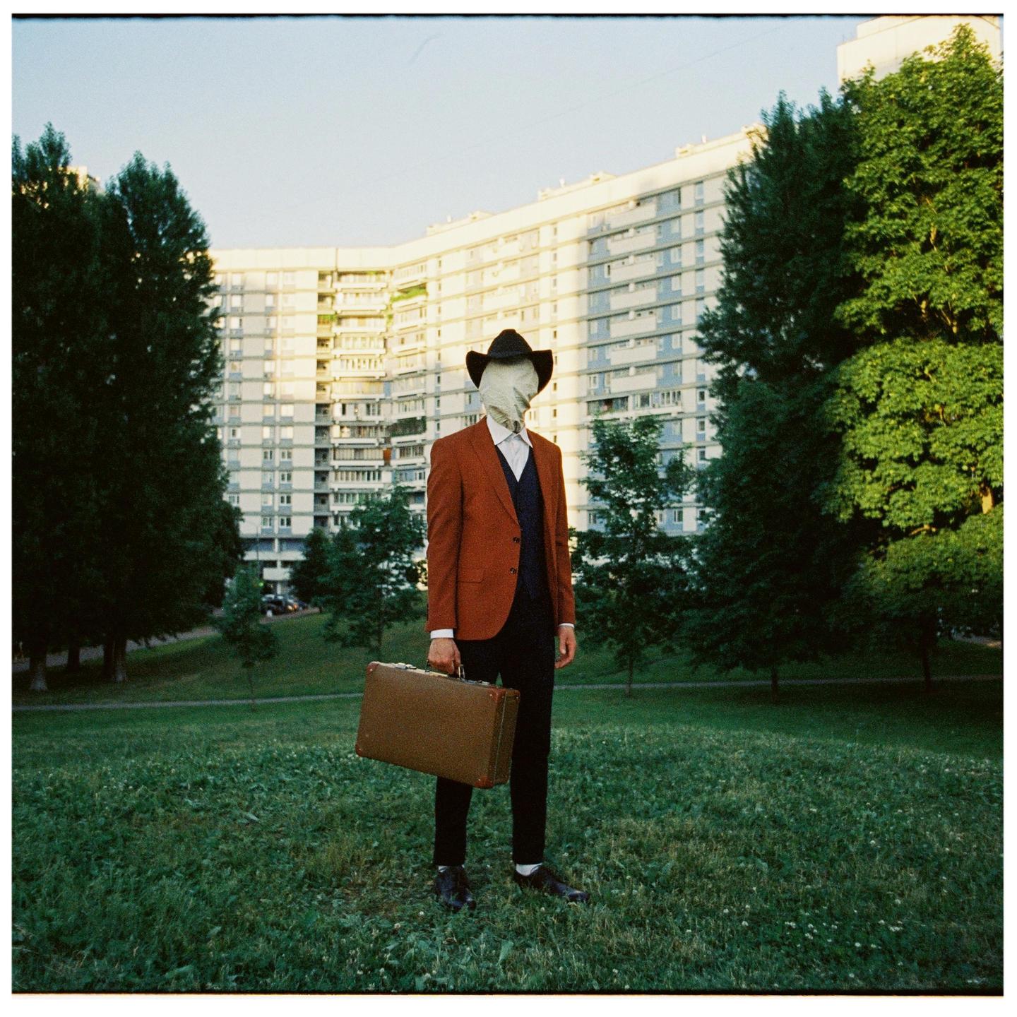 A person with a cloth covering their face and wearing a cowboy hat, suit jacket, and holding a briefcase stands in a grassy field with a large building and trees in the background.
