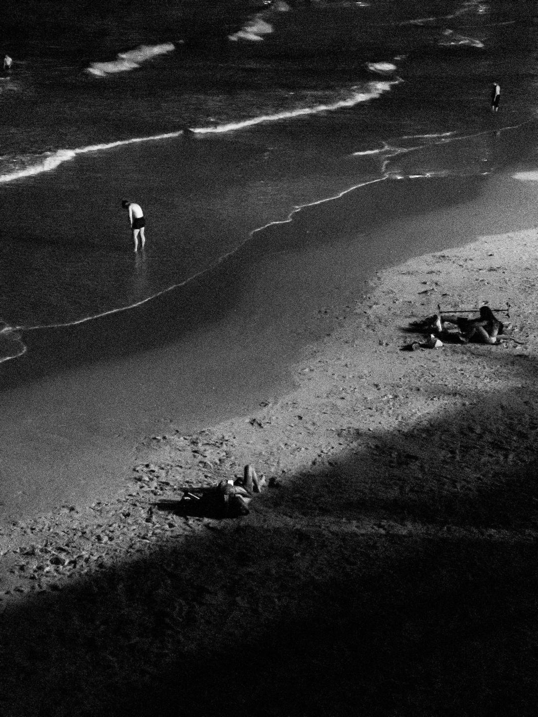 A person stands in the shallow water of a beach while two other people relax on the sand.