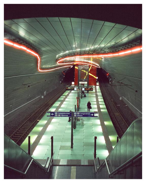 A person stands on a subway platform with light trails from a passing train overhead.