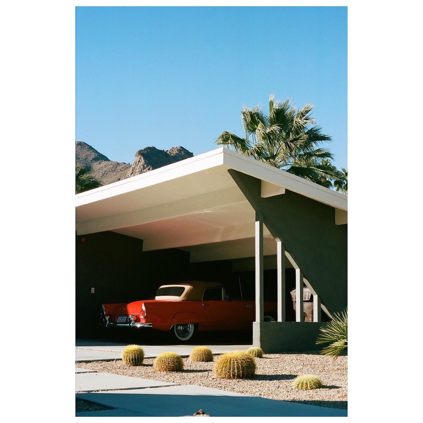 A red convertible car is parked under a carport with mountains and palm trees in the background.