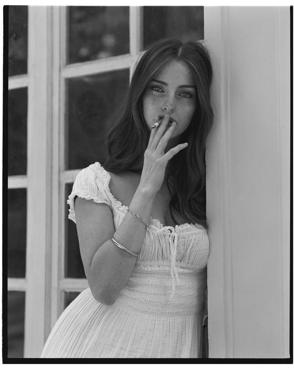 A woman with long hair and freckles leans against a wall and smokes a cigarette.