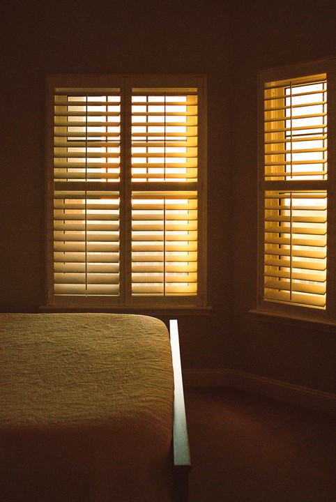 Sunlight streams through two windows with closed blinds next to a bed.