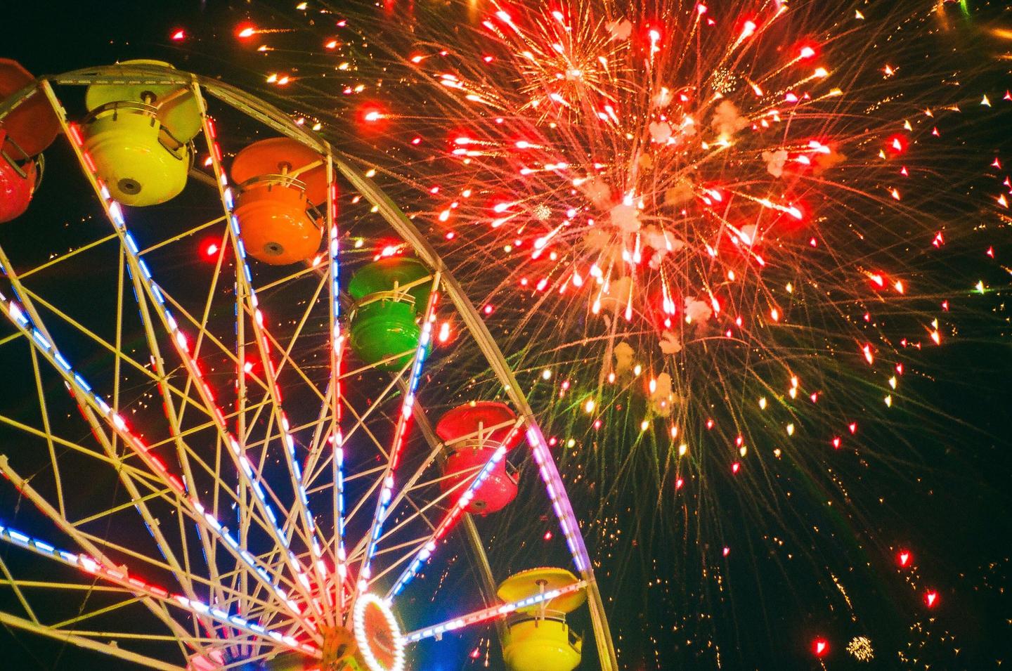 A ferris wheel with colorful gondolas is illuminated by fireworks exploding in the night sky.