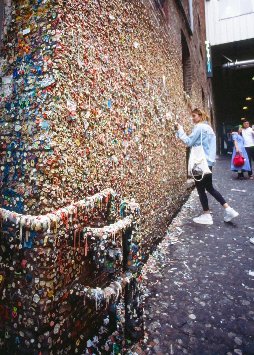 A woman stands next to a brick wall covered in colorful chewing gum.
