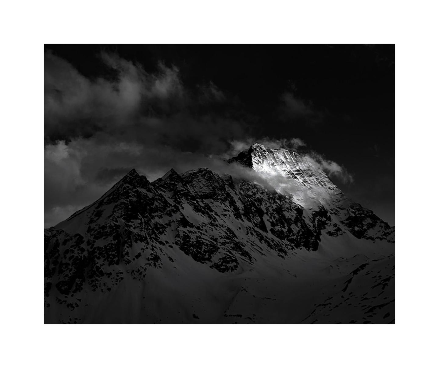 A snow-covered mountain peak is illuminated by a bright light against a dark, cloudy sky.