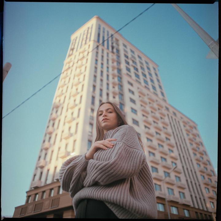 A woman in a chunky knit sweater stands in front of a tall building under a clear sky.