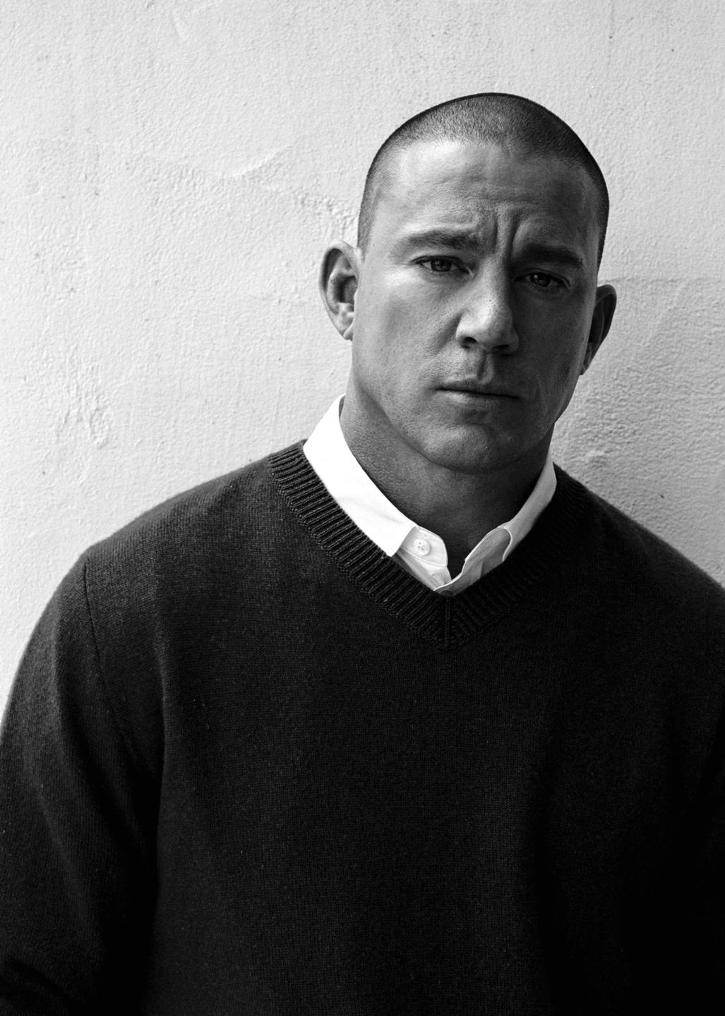 A man with a shaved head and a serious expression is wearing a sweater over a collared shirt.