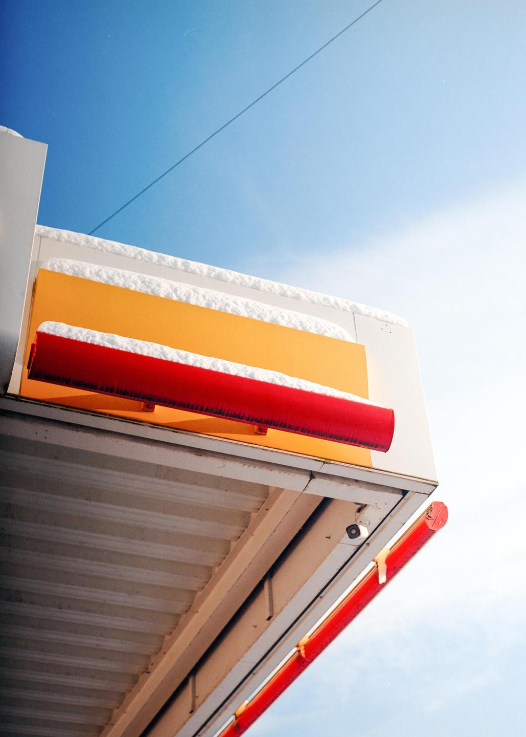 A gas station canopy with red and yellow accents is covered in snow under a bright blue sky.