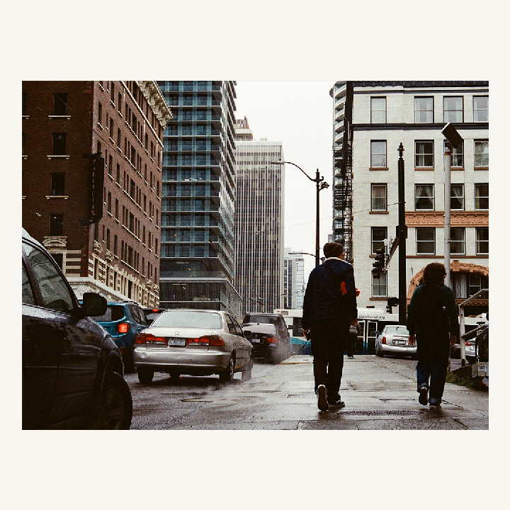 Two people walk away from the viewer on a wet city street with cars and tall buildings.