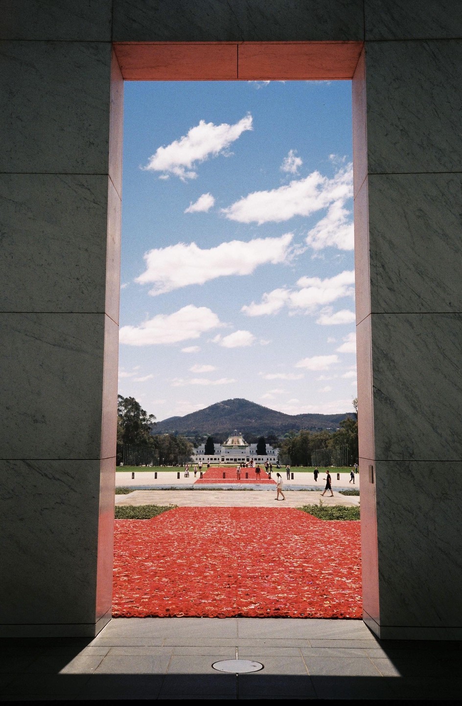 A view through a large rectangular opening shows a red poppy display leading to a distant building with a dome, a mountain, and a blue sky with clouds.