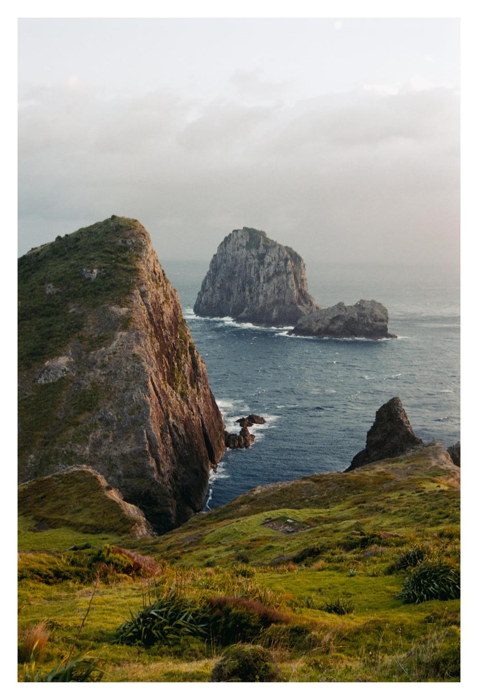 A dramatic landscape shows a steep, grassy cliff overlooking the ocean with several rocky islands in the distance under a cloudy sky.