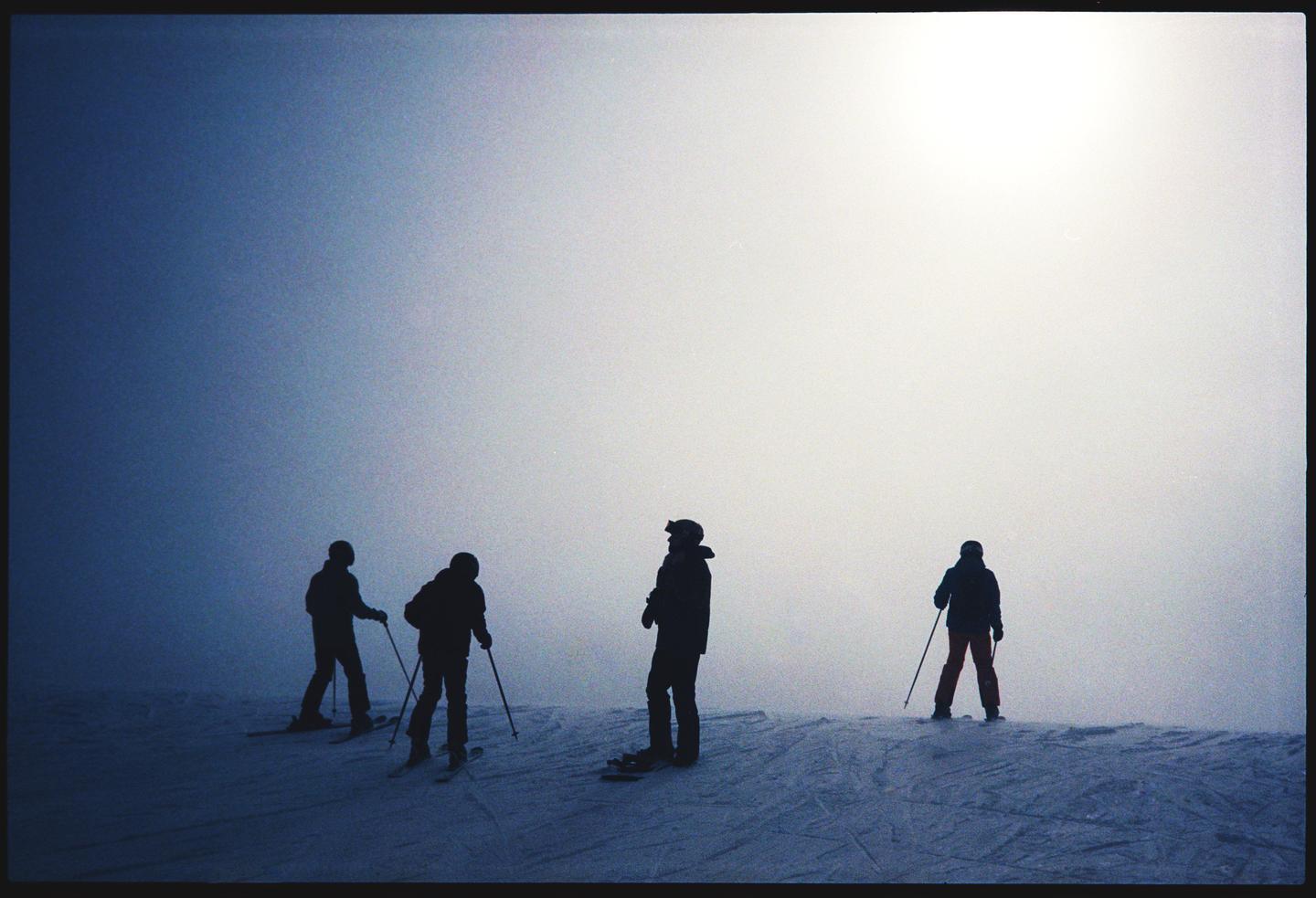 Four skiers are silhouetted against a bright, hazy sky on a snowy slope.