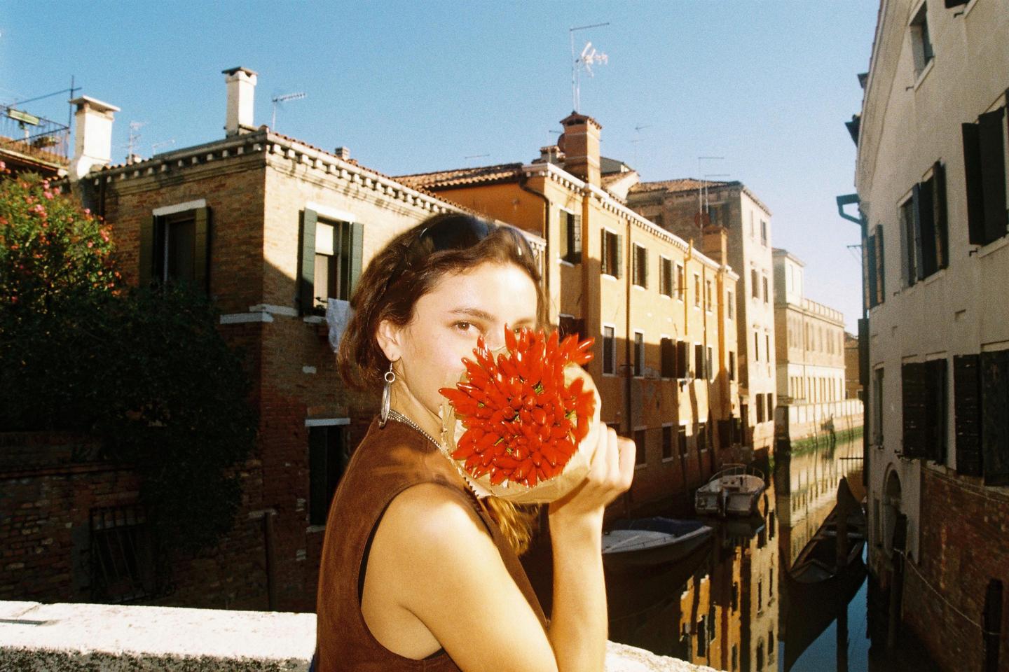 A woman holds a bouquet of red chili peppers in front of her face, standing on a bridge overlooking a canal in Venice.