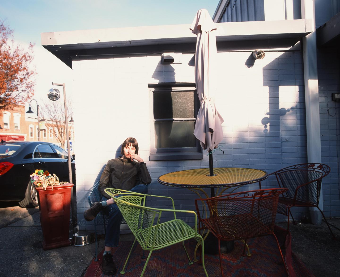 A woman sits in a green metal chair next to a table with an umbrella on a patio.