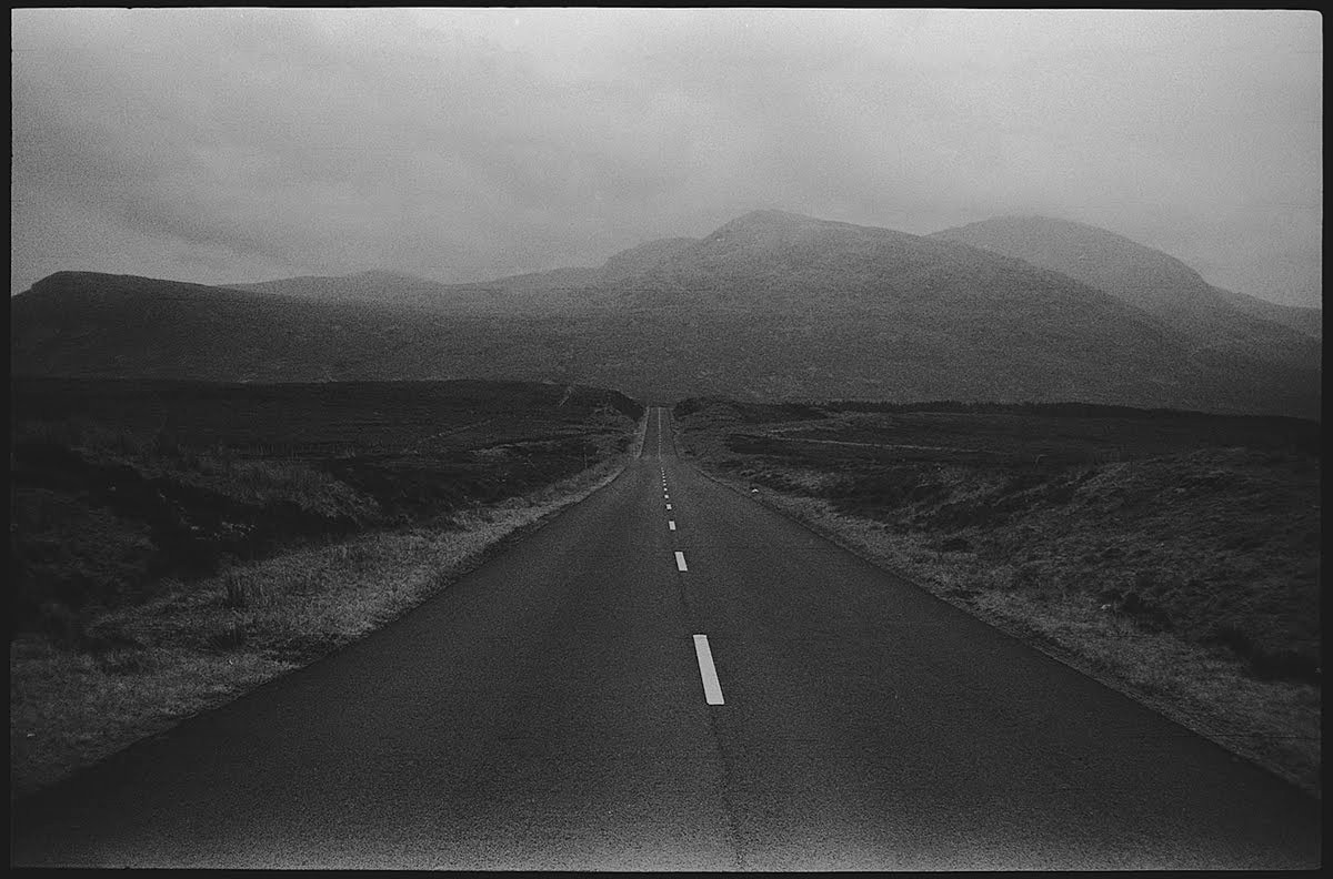 A long, straight road with a white dashed line down the center leads toward distant mountains under a hazy sky.