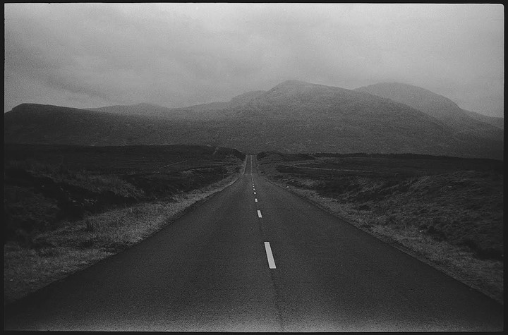 A long, straight road with a white dashed line down the center leads toward distant mountains under a hazy sky.