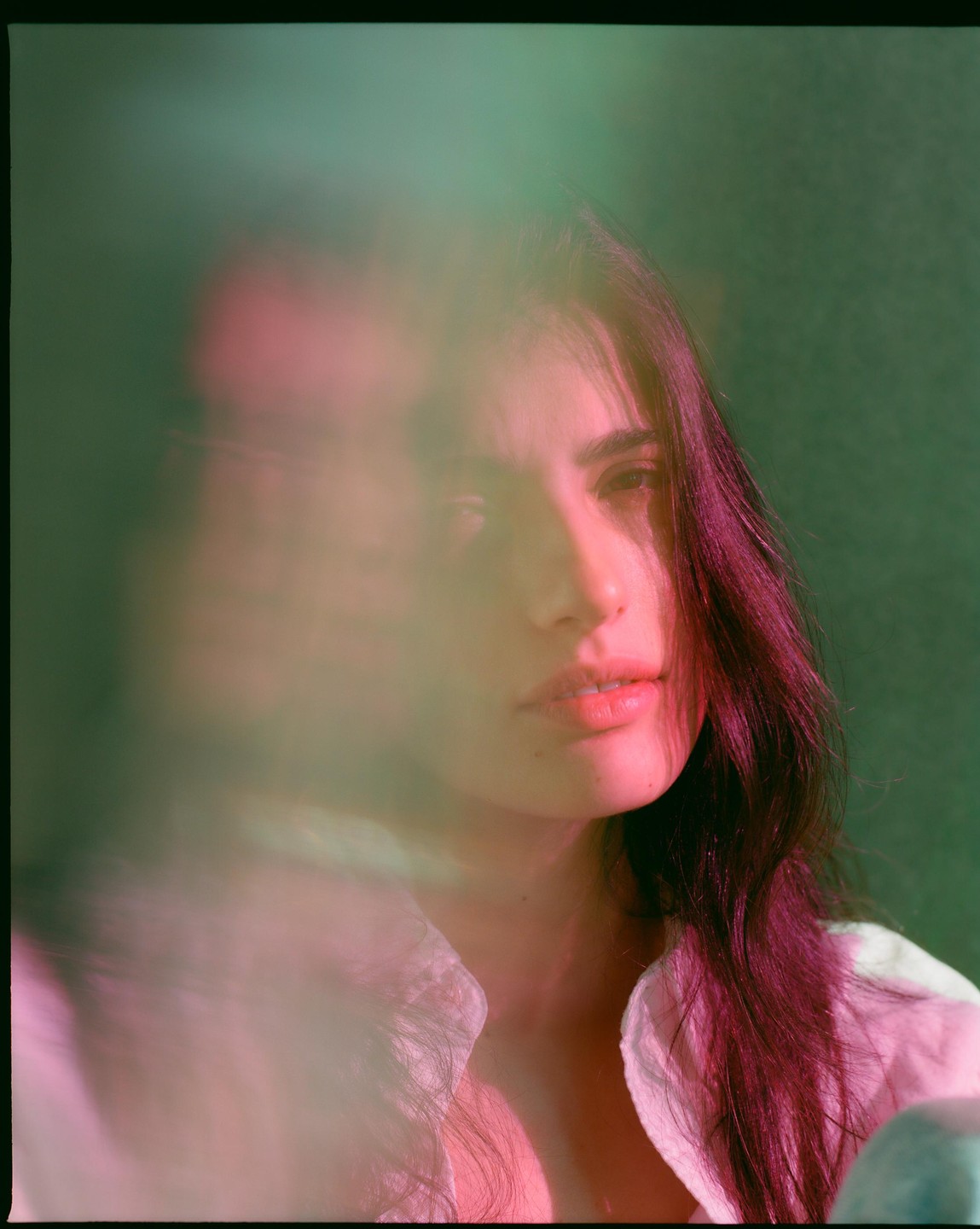 A woman with long dark hair looks directly forward, with a blurred pink and green foreground.