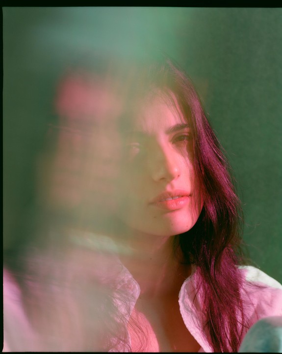 A woman with long dark hair looks directly forward, with a blurred pink and green foreground.