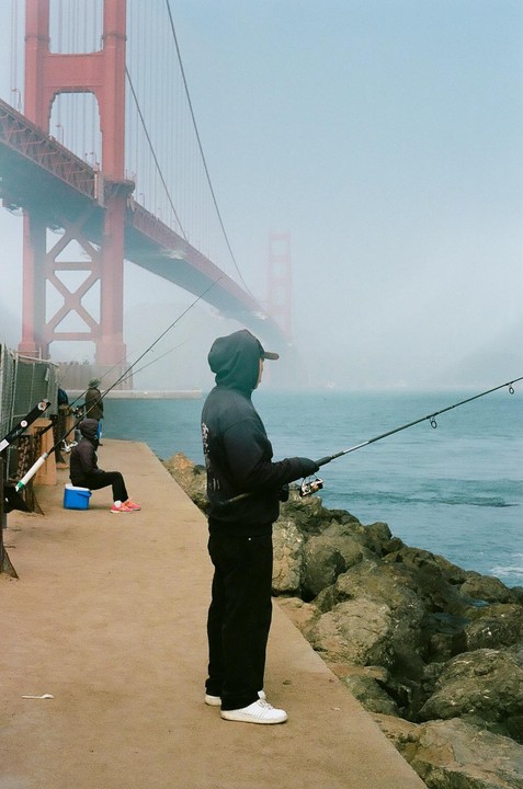 A man stands on a concrete pier fishing in the water with a bridge in the hazy background.