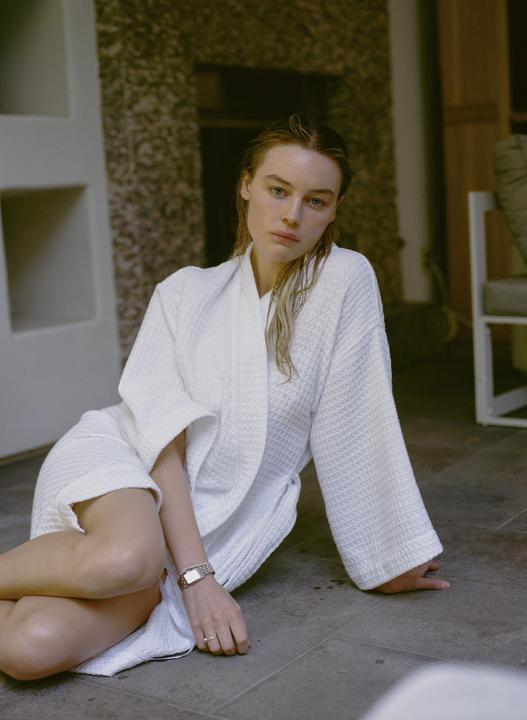 A woman in a white robe sits on a tiled floor looking directly at the viewer.