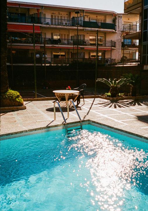 A swimming pool with sparkling water is in the foreground, with a table and chair next to it, and a building with balconies in the background.