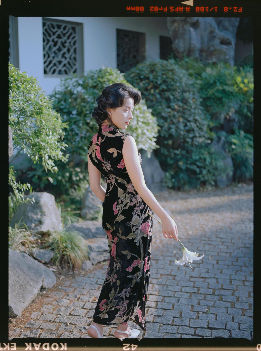 A woman in a floral dress holds a white lily in a garden with a building in the background.
