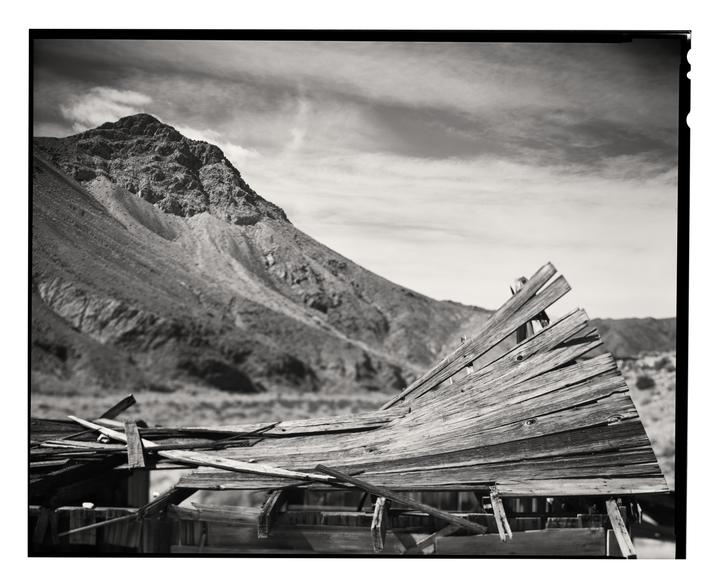 A dilapidated wooden structure sits in the foreground with a large mountain in the background under a cloudy sky.