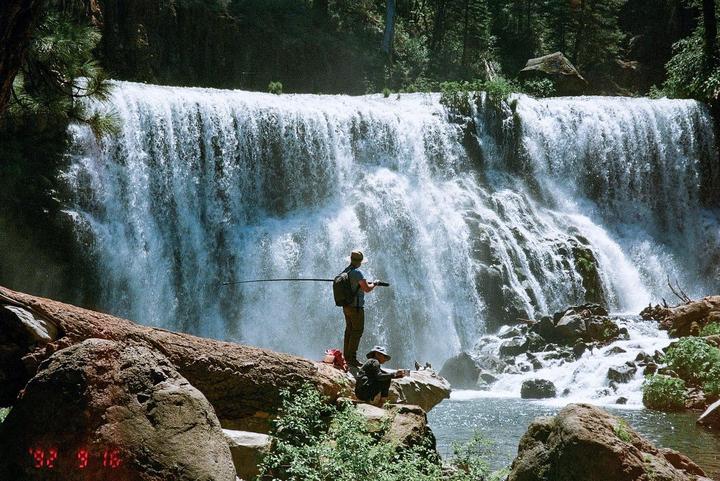 Two men fish in a river at the base of a large waterfall.