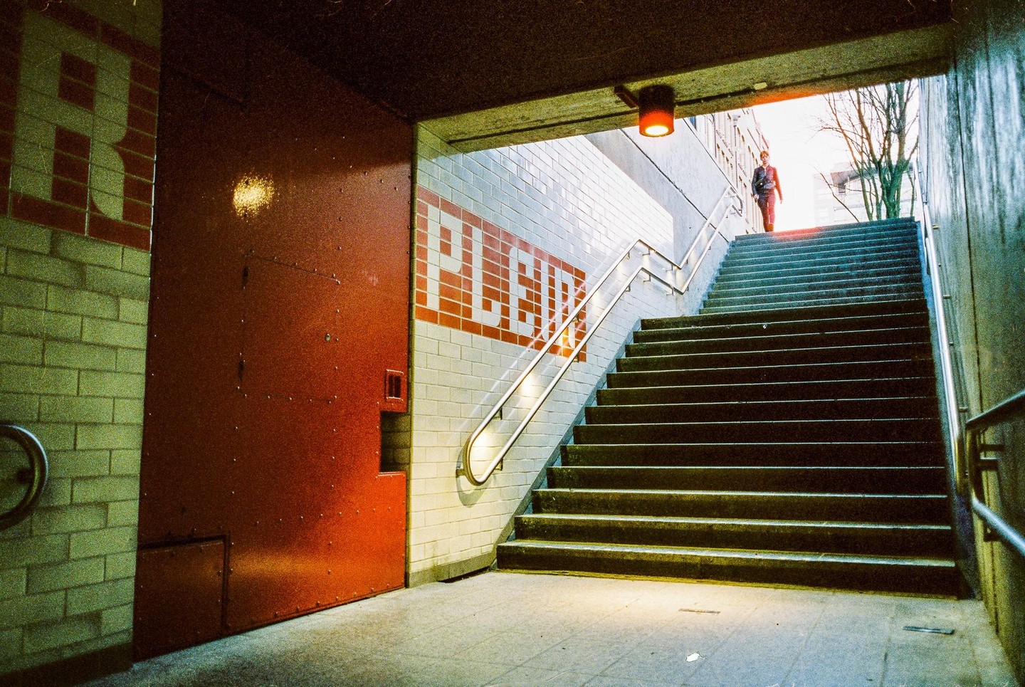 A person walks up a long staircase out of a subway station with a red door and tiled walls.