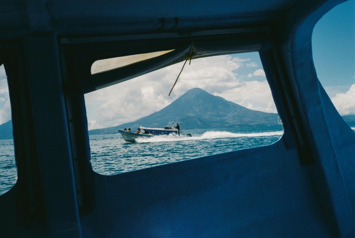 A boat with people on board speeds across a body of water with a large mountain in the background, viewed through a window.