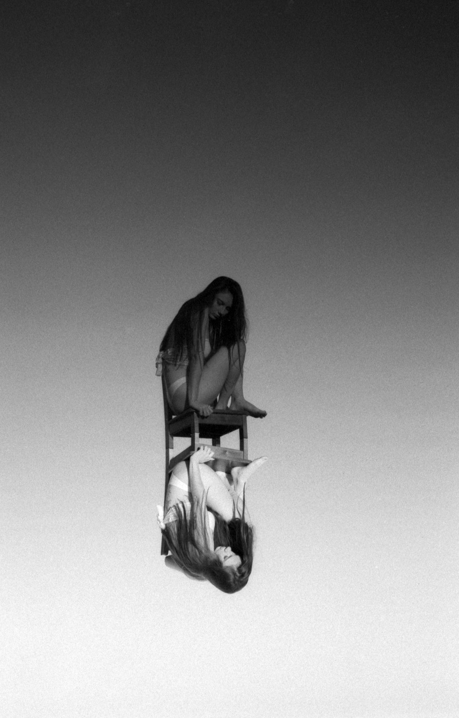 A woman sits on a chair with her reflection upside down below her, against a plain background.