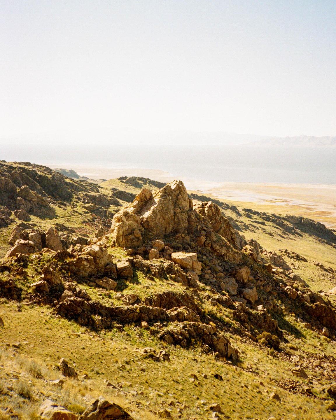 A rocky, grassy hillside overlooks a body of water and a distant shoreline.